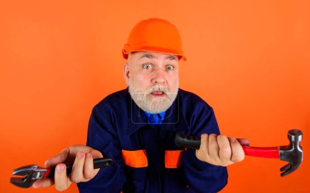 Confused builder in safety hard hat with adjustable wrench and hammer. Bearded mechanical worker or repairman in uniform and construction helmet with adjustable spanner and hammer. Tools for repair