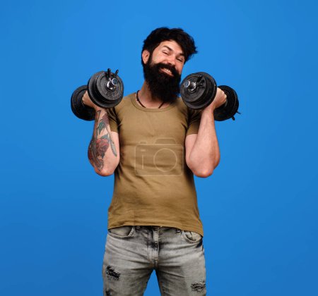 Photo for Strong handsome bodybuilder doing workout in fitness gym. Weight training. Sport equipment. Athletic muscular sportsman lifting dumbbells in sport club. Happy bearded man with dumbbells up in hands - Royalty Free Image
