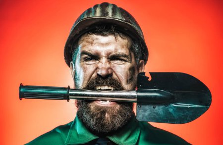 Photo for Mining, heavy industry and construction concept. Industrial worker, miner in safety hard hat with shovel in teeth. Angry bearded man in working uniform and helmet with sapper shovel or military spade - Royalty Free Image