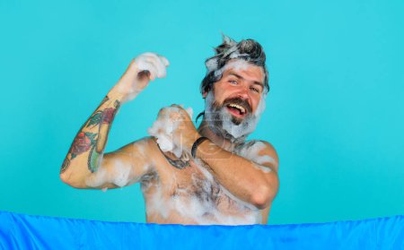 Hombre sonriente lavando el cuerpo con gel hidratante. Hombre guapo con el torso desnudo tomando ducha relajante en el baño. Cuidado corporal. Un tipo atractivo lavando el cuerpo con esponja de jabón. Hombre barbudo lavando el cabello