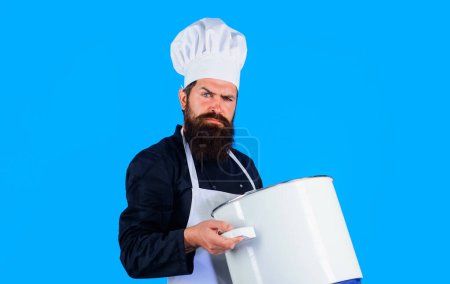 Kochen, Kulinarik, Lebensmittelzubereitung. Ernster bärtiger Mann mit Kochmütze und Uniform mit Auflauf oder Topf. Kochtöpfe, Geschirr, Besteck. Männlich kochen in Schürze mit großem Topf oder Pfanne. Küchenutensilien