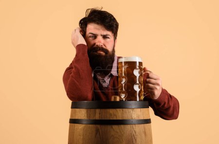 Beer time. Serious bearded man drinking draft beer in restaurant. Stylish handsome man with mug of beer on wooden barrel. Holiday, drinks, alcohol and leisure concept. Wooden barrel with craft beer