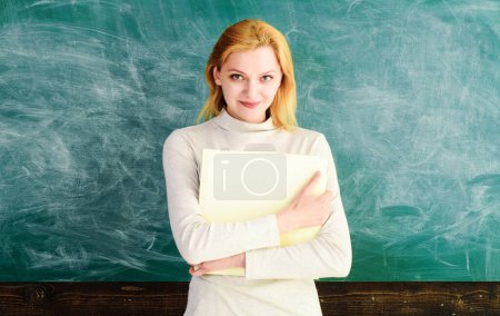 Smiling female teacher standing with teachers journal in front of chalkboard. School teacher, lecturer with class journal in classroom. Blonde student girl with book near blackboard in university