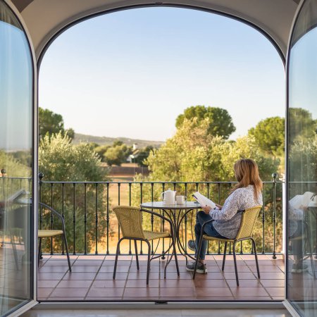 Téléchargez les photos : Femme lisant un livre sur la terrasse de sa chambre avec de grandes portes vitrées et vue sur l'extérieur d'un grand jardin - en image libre de droit