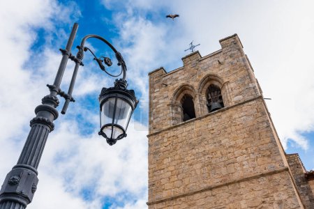 Mittelalterlicher Kirchturm und elektrische Straßenlaterne, wie ein Storch durch den Himmel fliegt. Aranda