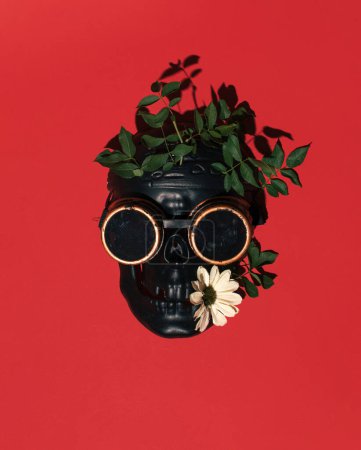 Photo for Black human skull mask decorated with greenery, flower and retro aviator goggles. Steampunk inspired layout against red background. - Royalty Free Image
