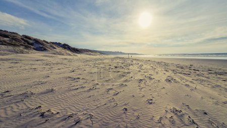 Foto de Los tonos cálidos del sol poniente arrojan un resplandor sereno sobre las dunas de la playa de Harlech en Gales, con las lejanas colinas creando una tranquila escena costera. Los patrones en la arena y la hierba marina en primer plano prestan una textura que habla a la calma - Imagen libre de derechos