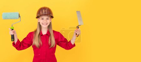 Téléchargez les photos : Une architecte. enfant adolescent souriant en chapeau dur constructeur avec rouleau de peinture et spatule sur le mur jaune. Bâtisseur d'enfants en conception d'affiche horizontale casque. En-tête de bannière, espace de copie - en image libre de droit