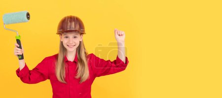 Téléchargez les photos : Heureux adolescent enfant dans constructeur casque dur avec rouleau de peinture sur le mur jaune. Bâtisseur d'enfants en conception d'affiche horizontale casque. En-tête de bannière, espace de copie - en image libre de droit