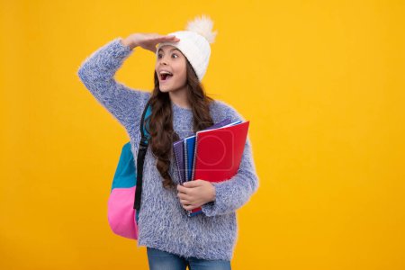 Téléchargez les photos : Portrait d'adolescentes en chapeau d'hiver portant des cahiers et un sac à dos sur fond jaune. L'école d'hiver. Visage excité, émotions joyeuses de l'adolescente - en image libre de droit