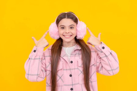 Téléchargez les photos : Happy girl enfant sourire pointant les doigts à chaud hiver fourrure oreille chauffe fond jaune, cache-oreilles. - en image libre de droit