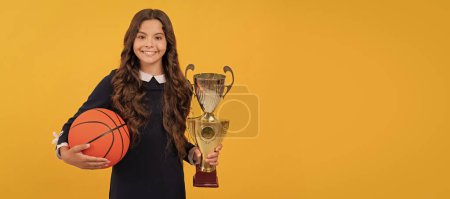 Téléchargez les photos : Cheerful kid hold basketball ball and champion cup on yellow background, sport success. Horizontal poster of isolated child face, banner header, copy space - en image libre de droit