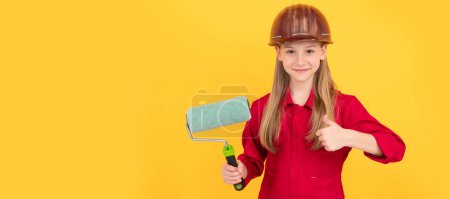 Téléchargez les photos : Rénovation enfant, adolescent enfant sourire dans le constructeur casque dur avec rouleau de peinture sur le mur jaune. pouce en l'air. Bâtisseur d'enfants en conception d'affiche horizontale casque. En-tête de bannière, espace de copie - en image libre de droit