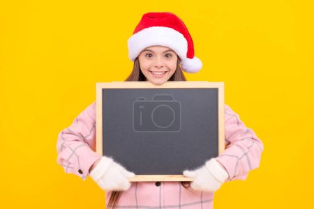 happy kid in santa claus hat hold blackboard for copy space. teen girl in mittens on yellow background. winter xmas holiday. child wearing warm clothes. back to school. happy new year. merry christmas Poster 626013546