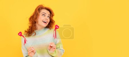 Téléchargez les photos : Portrait de femme isolé, bannière avec espace de copie. heureuse rousse fille avec rouge coeur bâtons sur fond jaune. amour - en image libre de droit