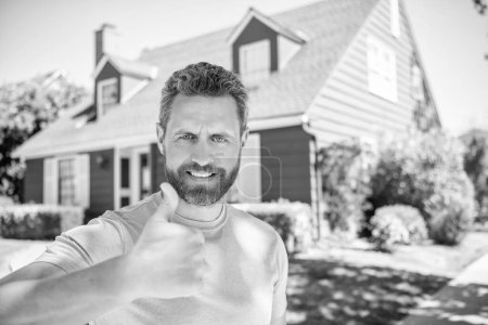 Téléchargez les photos : Heureux homme barbu montrant pouce sur fond de maison, banlieue. - en image libre de droit