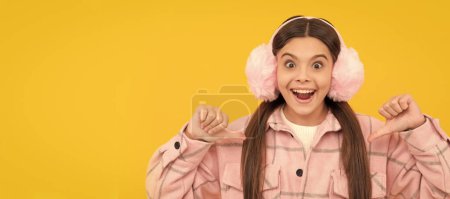Téléchargez les photos : Happy kid in warm winter fur ear warmers smile pointing thumb fingers at herself, fashion. Child face, horizontal poster, teenager girl isolated portrait, banner with copy space - en image libre de droit