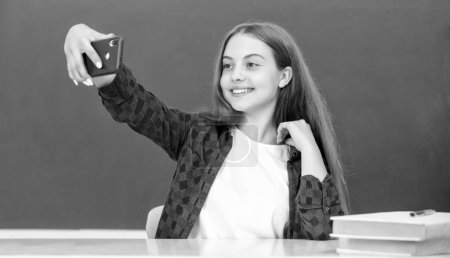 Foto de Niño feliz haciendo selfie en el teléfono inteligente en el aula en pizarra, retrato. - Imagen libre de derechos