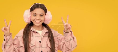 Téléchargez les photos : Happy girl child smile in warm winter fur earmuffs gesturing double V signs hand gesture. Child face, horizontal poster, teenager girl isolated portrait, banner with copy space - en image libre de droit