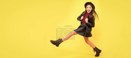 Foto de Happy teen girl in hat and leather clothes making wide step forward, step forward. Child face, horizontal poster, teenager girl isolated portrait, banner with copy space - Imagen libre de derechos