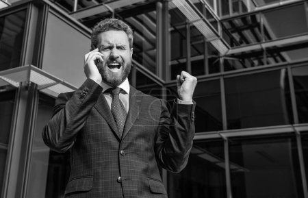Foto de Angry entrepreneur in businesslike suit shouting while phone conversation outside the office, frustration. - Imagen libre de derechos