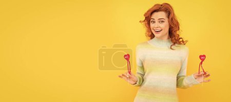 Téléchargez les photos : Portrait de femme isolé, bannière avec espace de copie. heureux rousse fille avec rouge coeur bâtons sur fond jaune - en image libre de droit