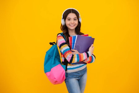 Back to school. Schoolgirl student in headphones with school bag backpack hold book on isolated studio background. School and education concept. Happy teenager, positive and smiling emotions of teen.