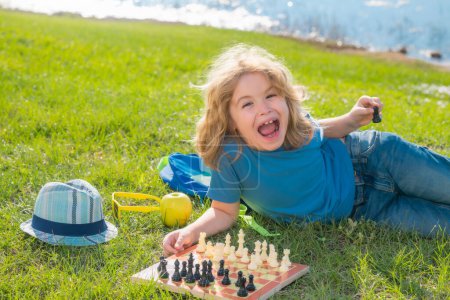 Concentrated kid developing chess strategy, playing board game in backyard, laying on grass. Logic game for kids and logical thinking