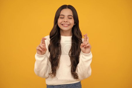 Pleased cheerful teenager child wishing good luck, crosses fingers, has faith for better, isolated over yellow background