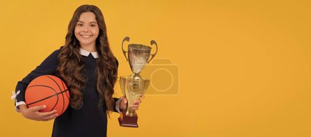 Téléchargez les photos : Glad child hold basketball ball and champion cup on yellow background, victory. Horizontal poster of isolated child face, banner header, copy space - en image libre de droit