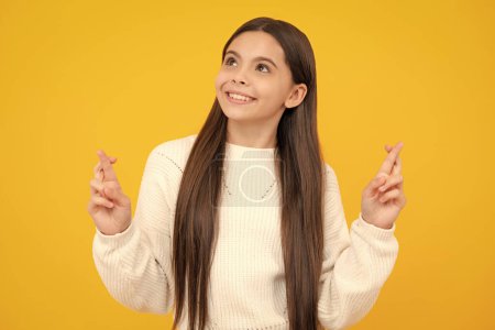 Glad teenager girl crosses fingers, anticipate hearing good news, isolated yellow background. Happy child rejoices that his dreams came true. Excitement concept