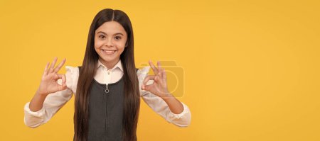 Happy girl child in school uniform smile gesturing double OK sings yellow background, okay. Child face, horizontal poster, teenager girl isolated portrait, banner with copy space