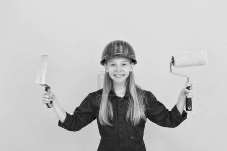 Photo for Cheerful teen child in builder hard hat with paint roller and spatula on yellow wall. - Royalty Free Image