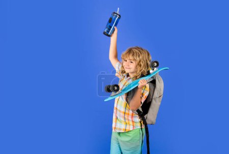 Child boy holding longboard and water bottle on blue isolated background. Kid with pennyboard. Studio shot of cheerful little fashion kid with penny board