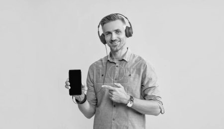 smiling mature man listen music in headphones pointing finger on phone, presentation.