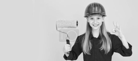 Builder teenager girl in helmet. happy teen child in builder hard hat with paint roller on yellow wall. ok. Child builder in helmet horizontal poster design. Banner header, copy space