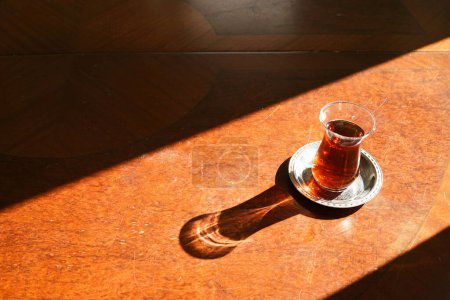 Foto de Un vaso de té turco en la mesa de madera con el sol brillando en ella - vaso de té turco especial y decorado pequeño plato de metal - tradición de Turquía - Imagen libre de derechos