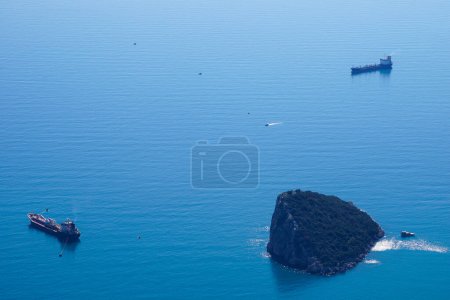 The cargo ships are at anchor near Antalya (Turkey) bay with the rocky island in the clean blue water - aerial drone view