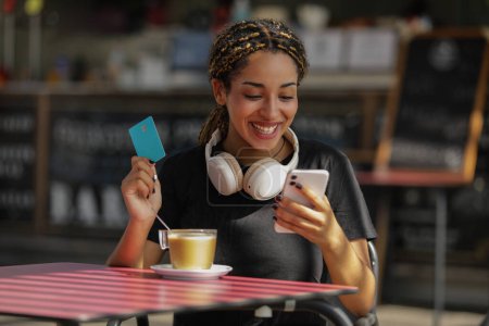 Foto de Mujer sonriente usando una tarjeta de crédito y un teléfono móvil mientras está sentada en una terraza del bar - Imagen libre de derechos