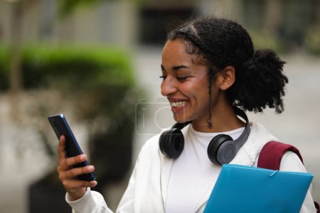 Foto de Sonriente joven estudiante mirando su teléfono inteligente al aire libre - Imagen libre de derechos
