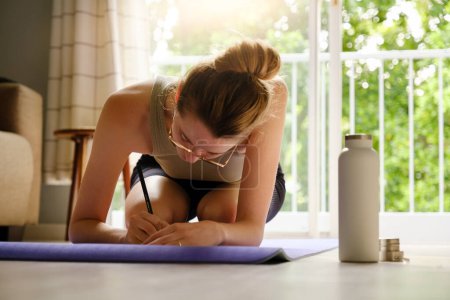 Mujer caucásica joven haciendo una entrada de diario mientras está sentado en una esterilla de yoga en casa con botella de agua de pie junto a la esterilla. Concepto de salud y bienestar.