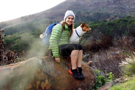 Téléchargez les photos : Portrait d'une femme caucasienne souriante en vêtements d'hiver assise avec son chien sur un sentier de montagne tôt le matin - en image libre de droit