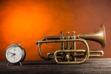 Photo for Brass trumpet on wooden table, orange background with antique clock - Royalty Free Image