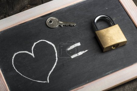 Photo for Blackboard with a drawing of a heart equal sign and a real padlock pogiato above, isolated on a wooden table - Royalty Free Image