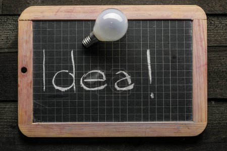 Photo for Blackboard with the inscription "idea!" and a light bulb resting on top, isolated on a wooden table - Royalty Free Image