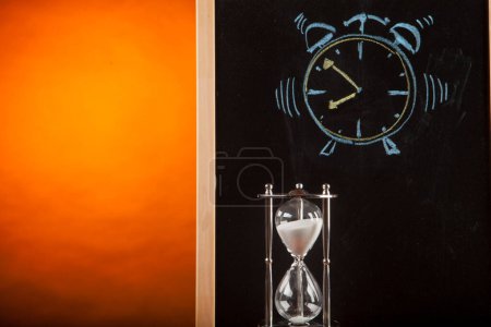 Photo for Alarm clock drawn with chalk on blackboard together with real hourglass isolated on orange background - Royalty Free Image