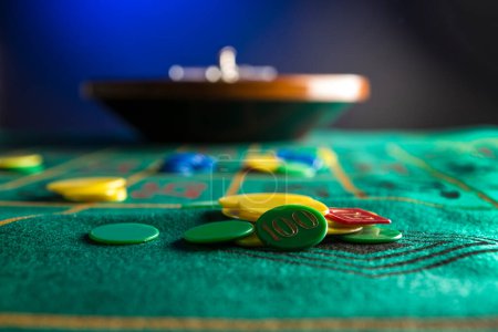 Foto de Rueda de ruleta de madera aislada sobre una mesa de juego verde con fichas de colores, iluminada por una luz azul - Imagen libre de derechos