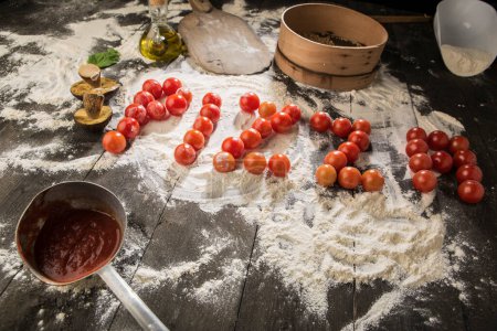 Foto de Escrito "pizza" con tomates cherry en una mesa llena de ingredientes como harina, salsa de tomate, aceite, champiñones, etc.. - Imagen libre de derechos