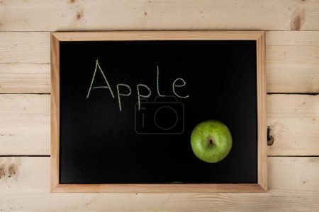 Photo for School blackboard on wooden table with "Apple " written on it and a real green apple - Royalty Free Image