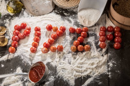Téléchargez les photos : Pizza écrite avec des tomates cerises sur une table pleine d'ingrédients tels que farine, sauce tomate, huile, champignons, etc.. - en image libre de droit
